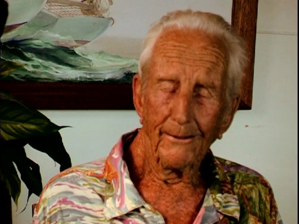 An elderly man recounts personal memories in an interview setting, interspersed with contemplative visuals of a woman standing by the ocean at sunset.