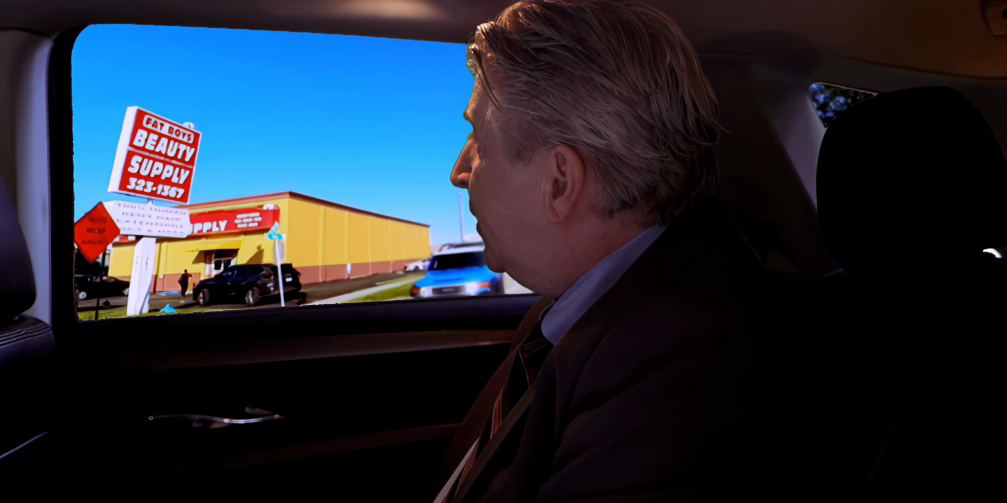 Two men travel in the backseat of a vehicle while discussing their surroundings and engaging in a professional conversation. They observe local commercial businesses through the window as they pass by.
