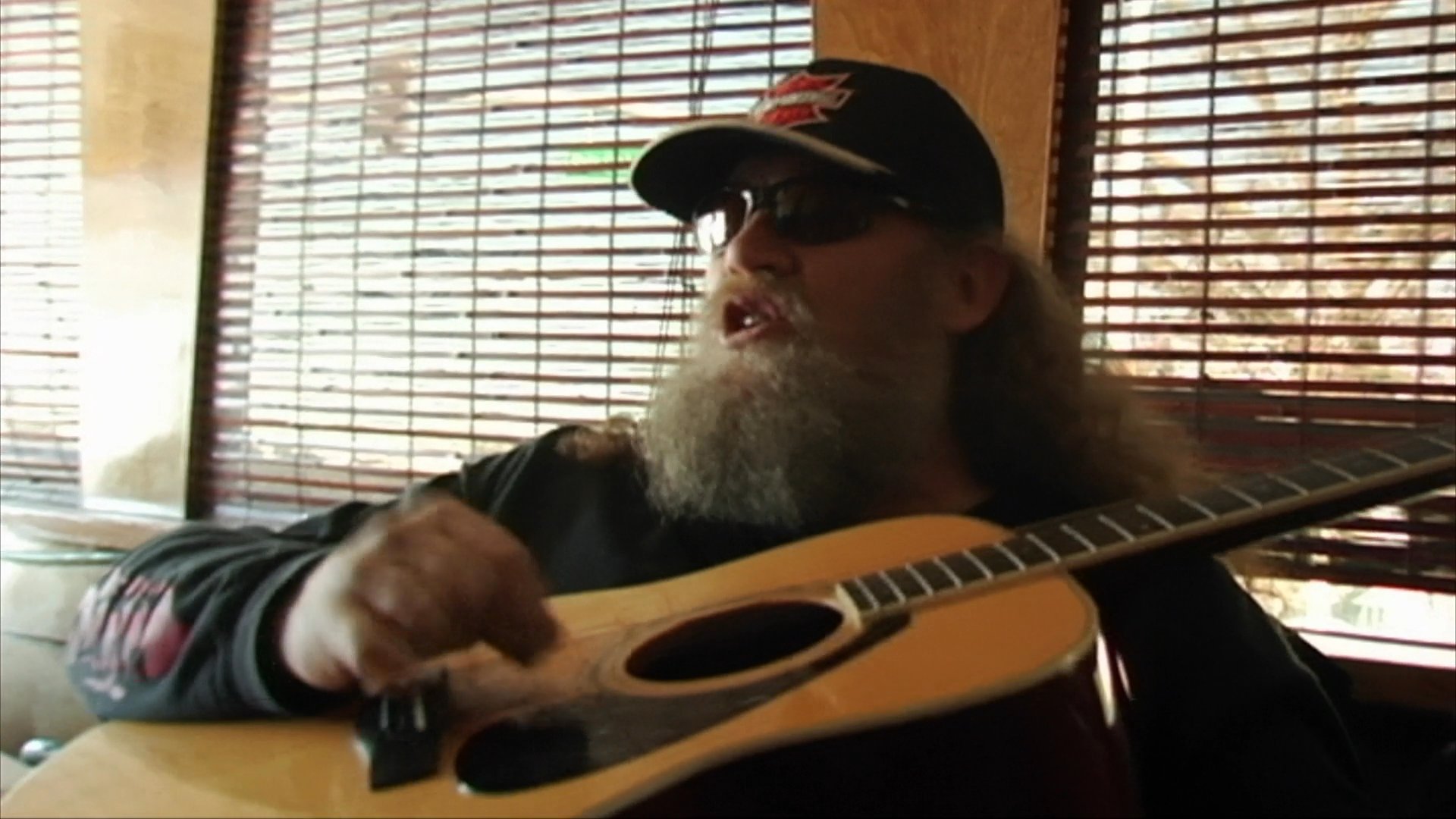 A bearded musician in a cap performs on an acoustic guitar while singing. The performance transitions from an outdoor setting to an indoor environment with blinds in the background.
