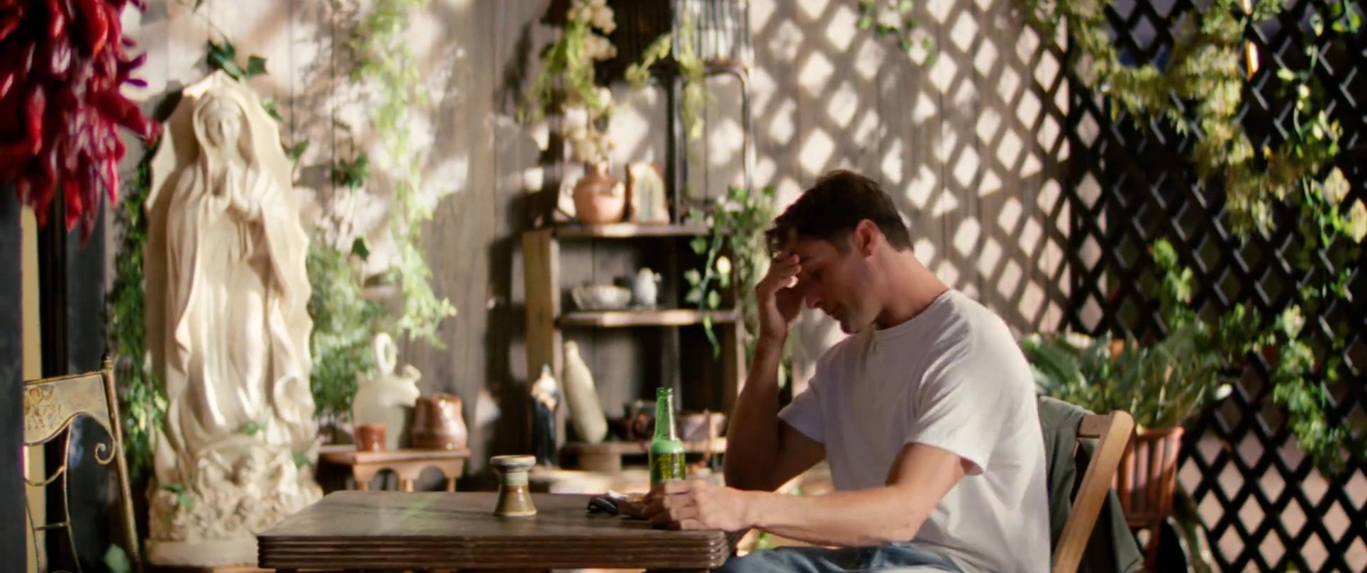 Two men exchange words in an outdoor, rustic patio environment decorated with dried chili peppers and greenery. The interaction concludes with one of the men standing and departing the table.