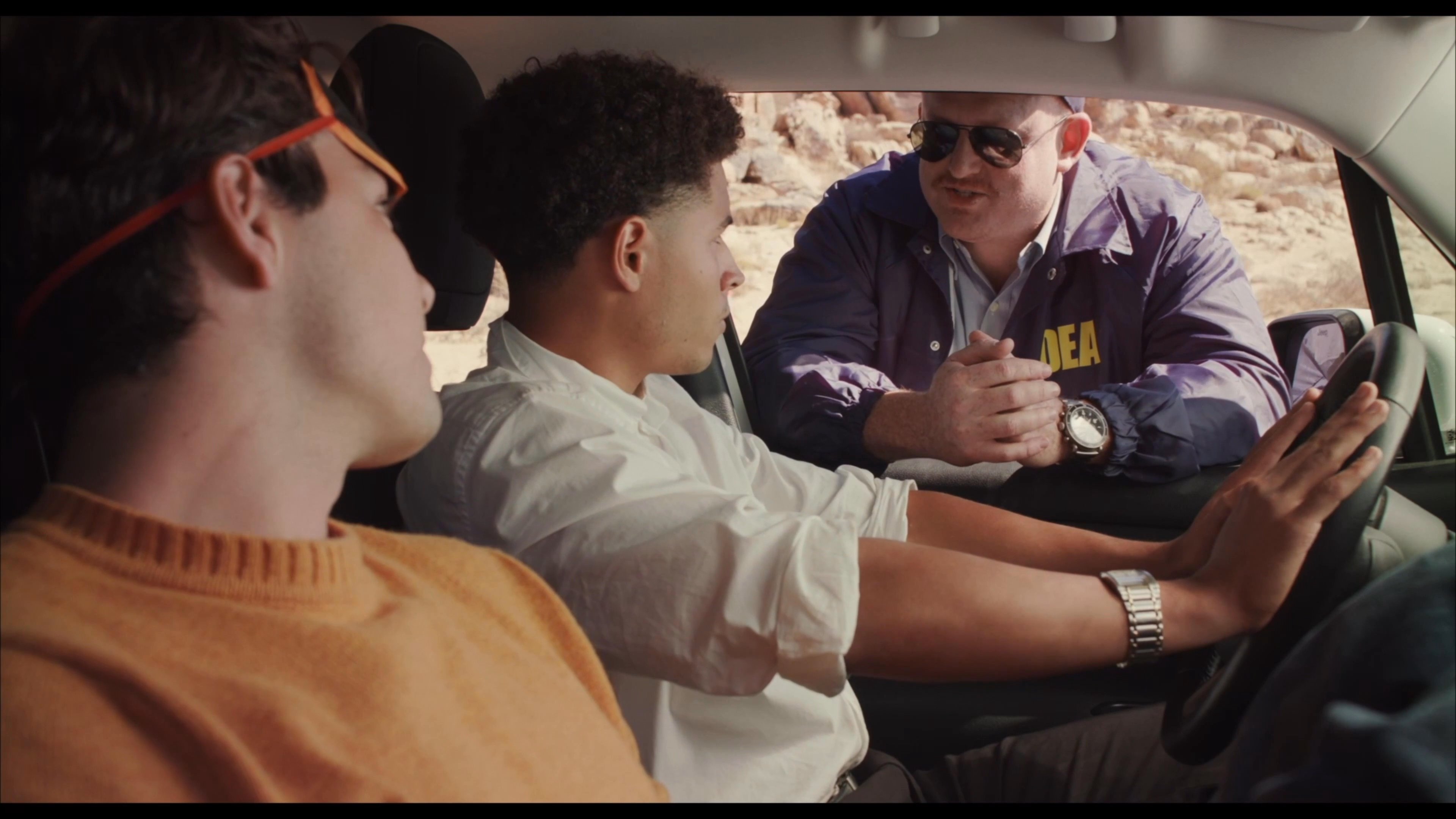 An agent wearing a DEA windbreaker conducts an interrogation from outside a vehicle, questioning two men seated inside. The interaction takes place in a remote desert environment.