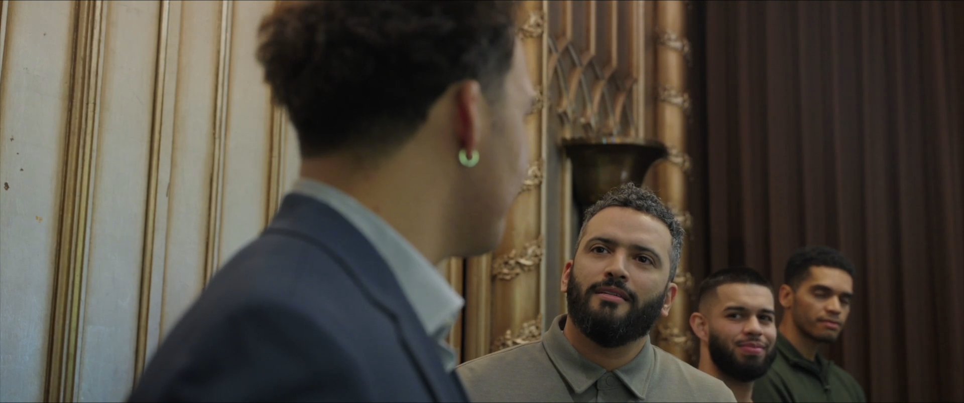 Two men engage in a professional conversation within an elegantly designed room, while other individuals stand in the background. The interaction focuses on interpersonal exchange in a formal social setting.