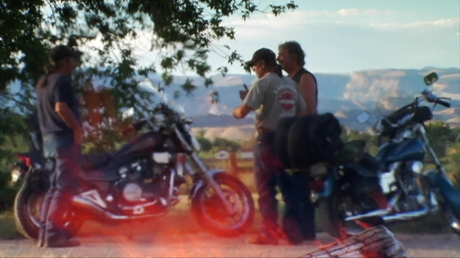 A group of motorcyclists pauses to gather near their parked bikes against a backdrop of desert terrain. They eventually continue their journey, riding past a rural structure.