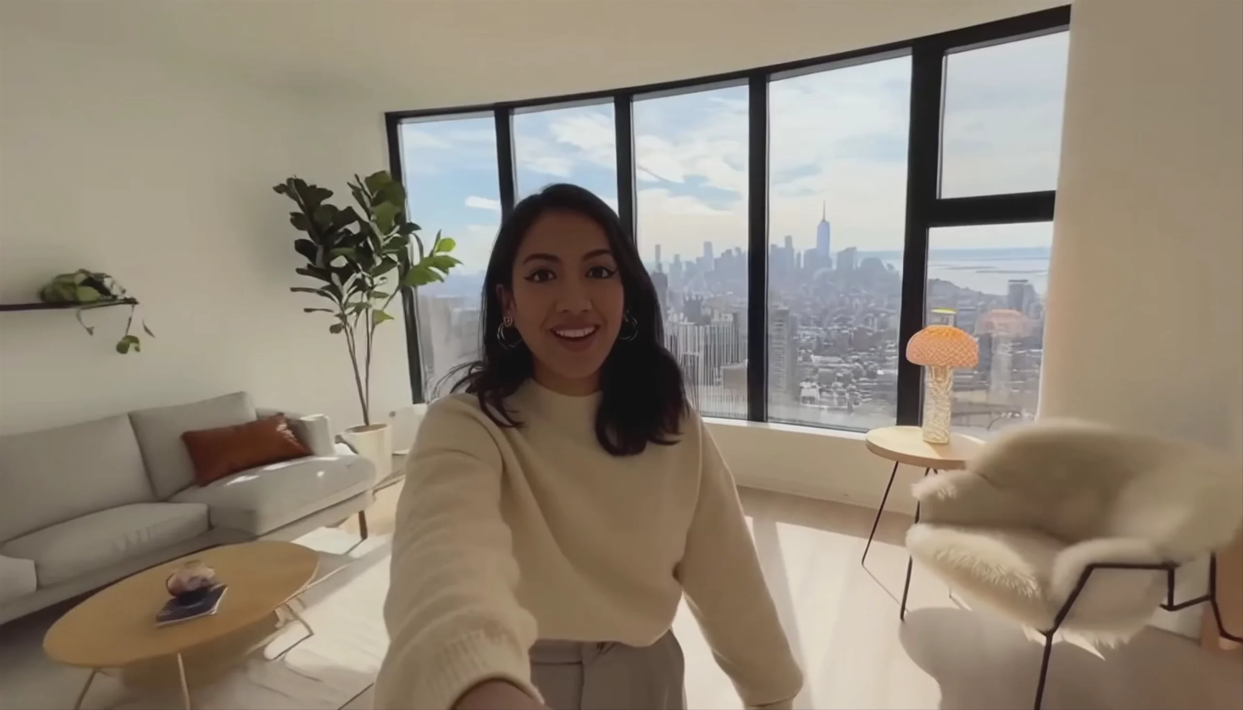 A first-person vlog-style video frame generated by Sora 2 Pro at 1792x1024 resolution. A smiling woman in a cream sweater stands in a bright, minimalist high-rise apartment. Behind her, large floor-to-ceiling windows reveal a sprawling city skyline under a clear blue sky, next to a modern sofa and chair.