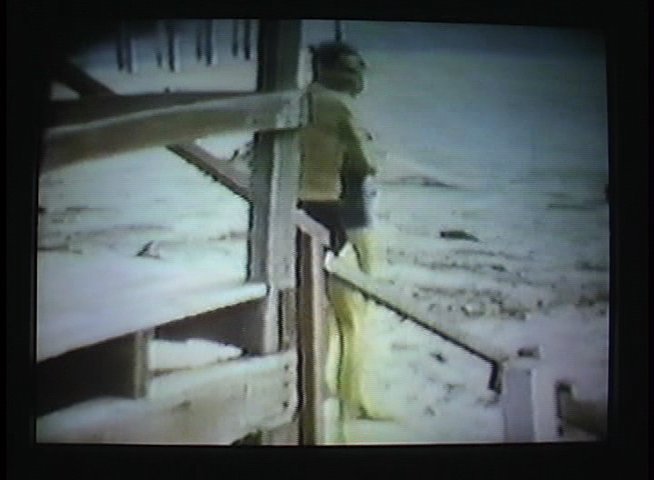 An elderly man wearing headphones watches archival film footage of people vacationing at a beach, reflecting on past memories.