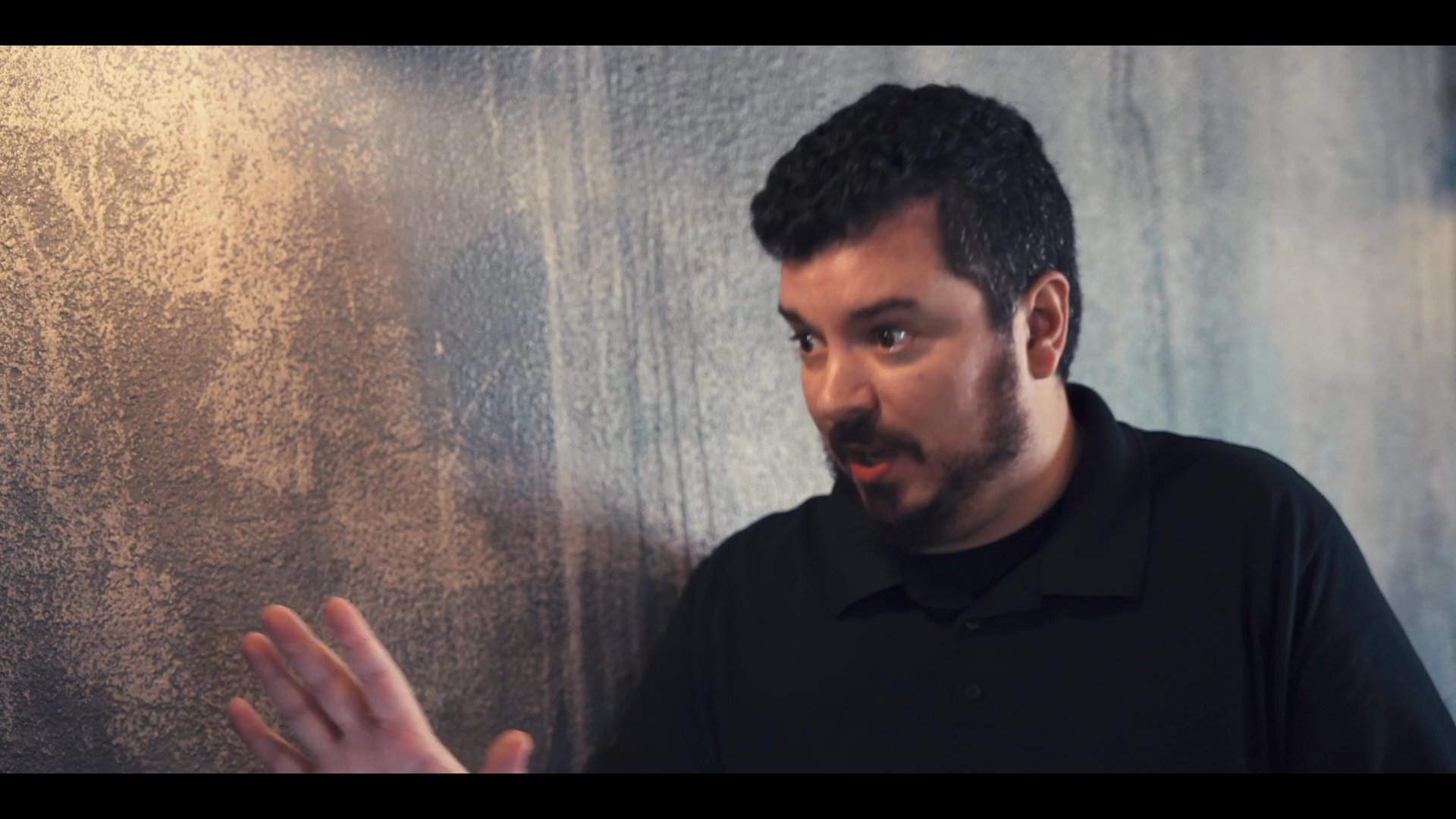 A man in a black polo shirt actively speaks and uses hand gestures to communicate, suggesting an instructional or persuasive dialogue.