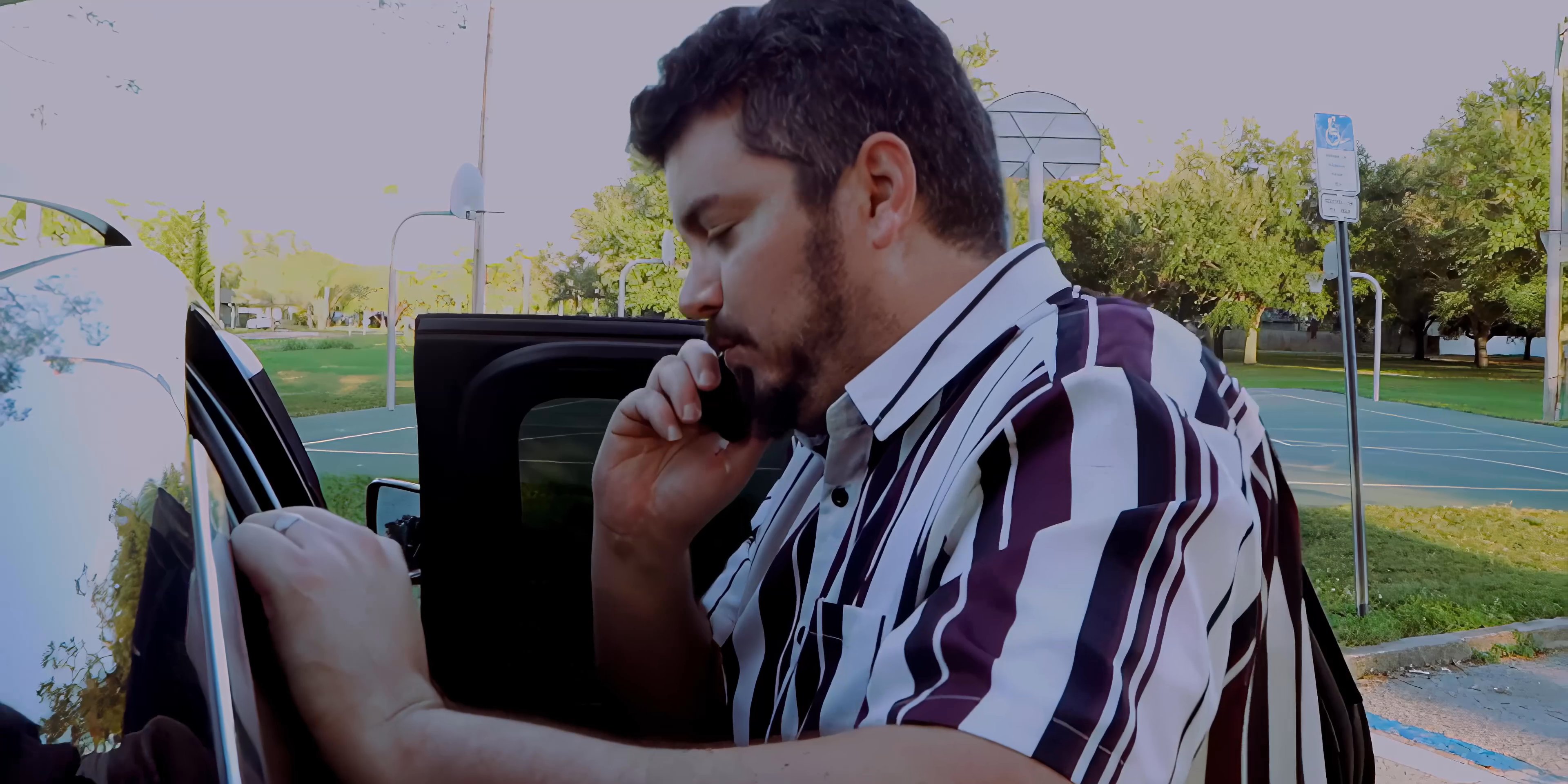 A man in a striped shirt stands by his open car door in a parking lot while engaged in a mobile phone conversation.