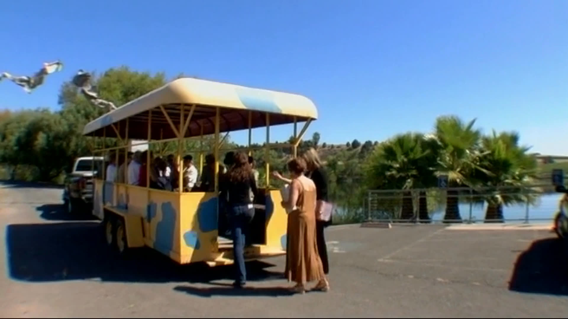 An elderly man discusses sharing art with a broader audience. Visitors ride an open-sided shuttle through a waterside venue, viewing colorful animal sculptures and an art display.
