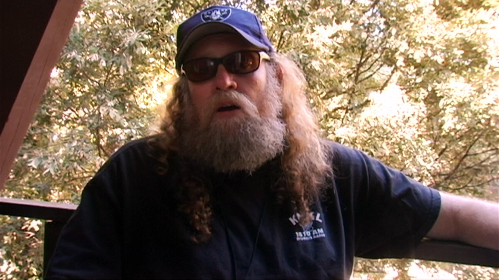 A man with a white beard and cap speaks directly to the camera while situated in a natural, wooded setting during an interview.