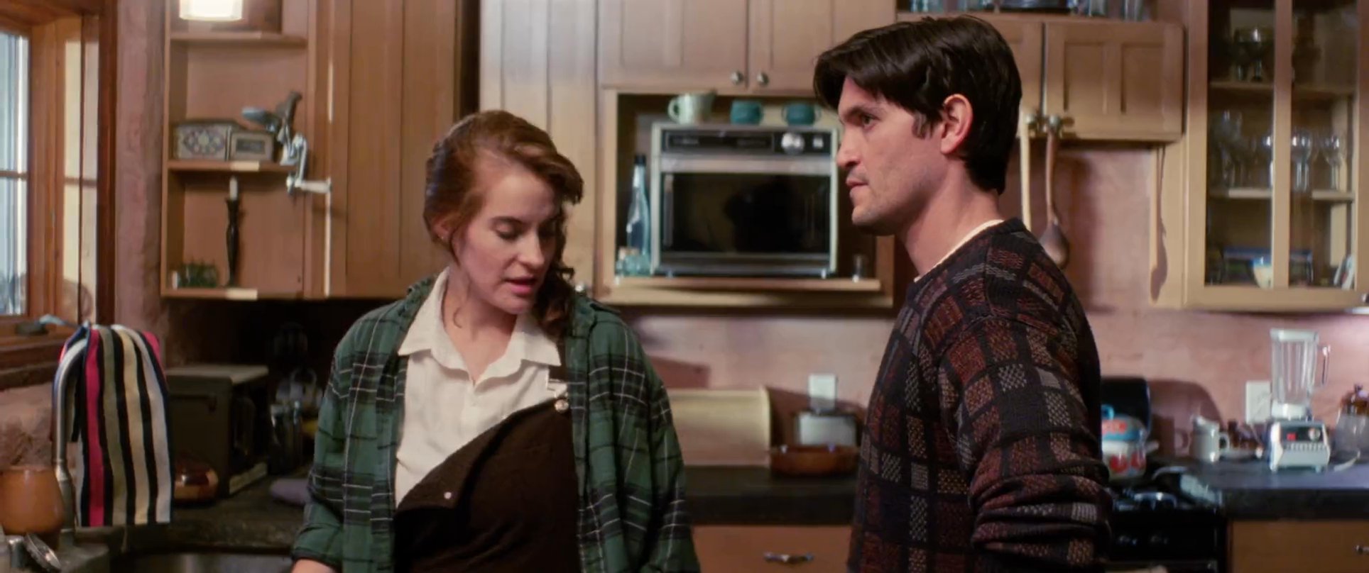 A man and woman engage in a tense conversation within a residential kitchen before ultimately shaking hands. The interaction shifts from a confrontational exchange to a moment of mutual understanding.
