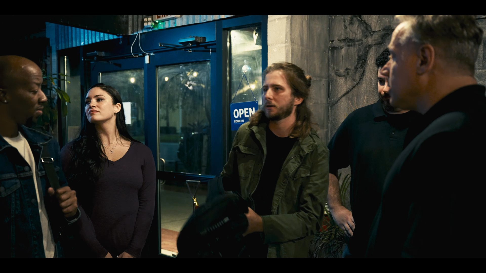 A diverse group of individuals engages in an animated conversation outside a building entrance at night. They exchange gestures and discuss matters in a casual, yet focused interaction.