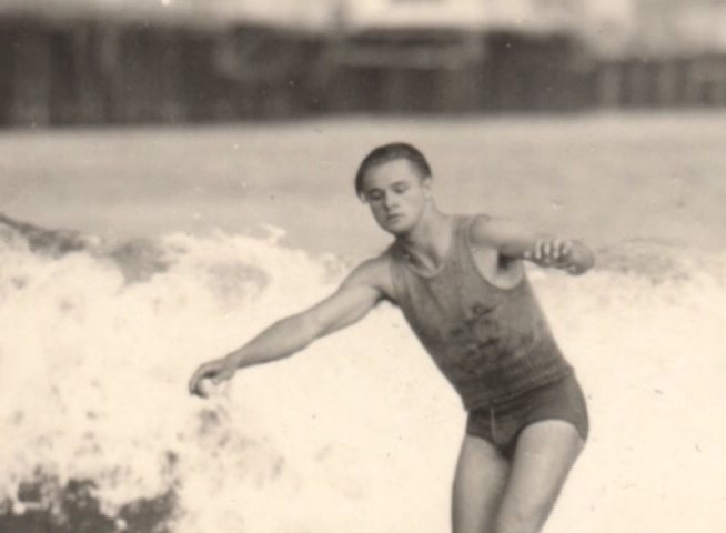 An elderly man discusses his past while archival, sepia-toned footage displays a young surfer riding ocean waves.