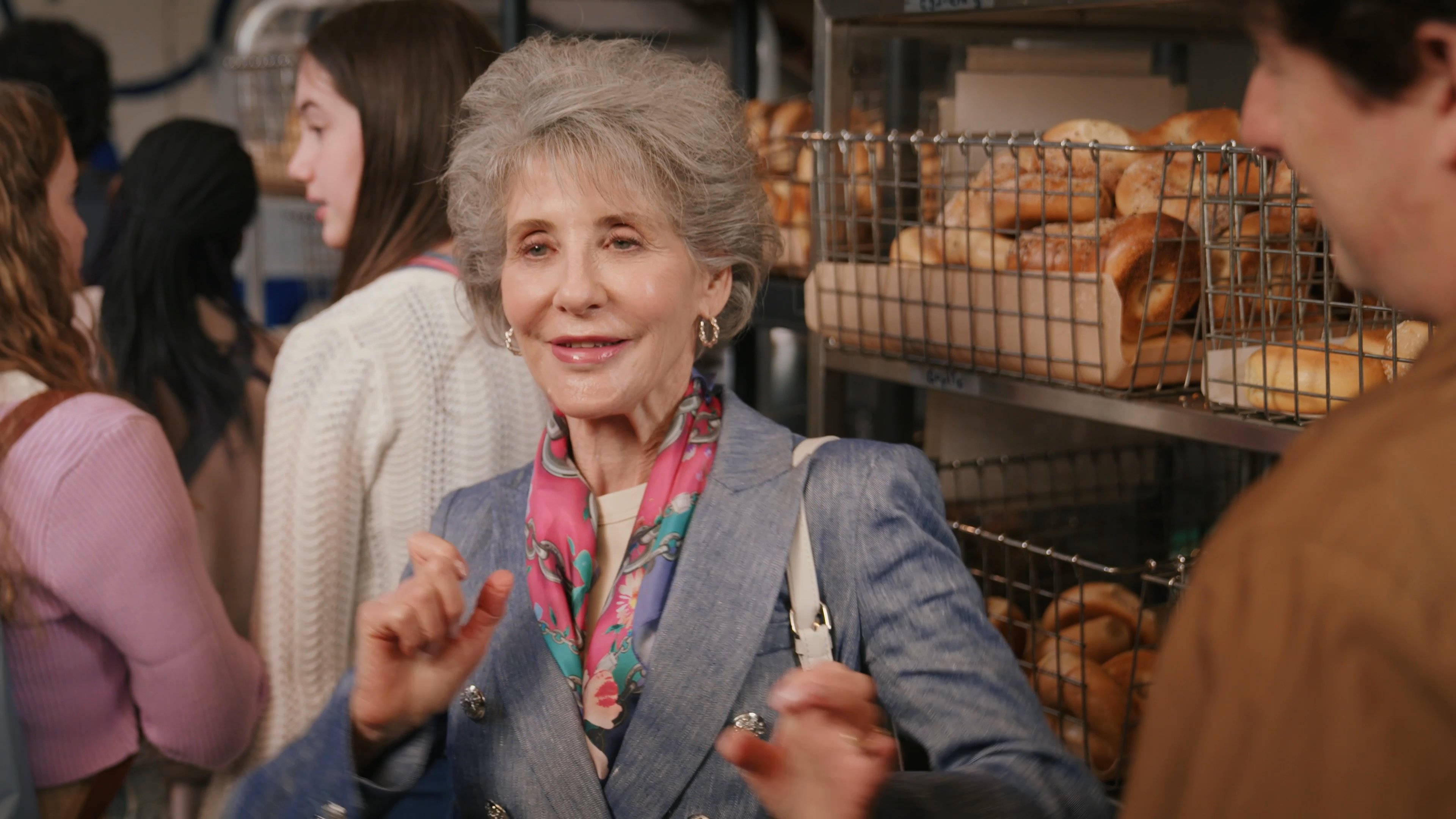 An older woman engages in an enthusiastic conversation with people inside a bustling bakery. She gestures expressively while speaking to a group standing near the product display cases.