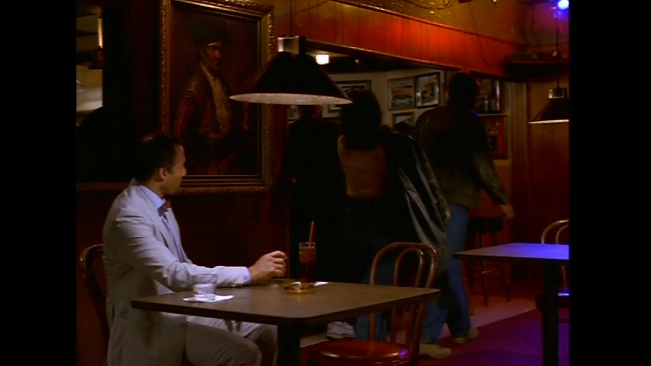 Three individuals converse and interact at a table inside a dimly lit diner. The scene captures the shift in social dynamics as participants depart and others remain.