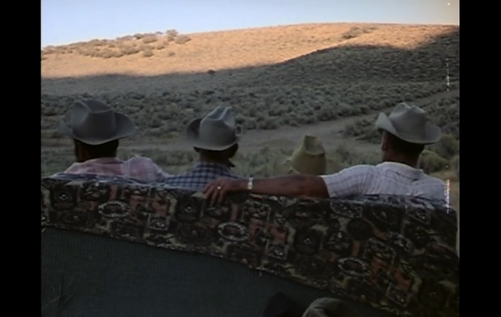A ranch gate opens onto a remote cow camp amid dry hills and sagebrush, where cattle graze and dust rises across the range. Cowboys watch the landscape from a wagon or bench as smoke drifts from a nearby cabin.