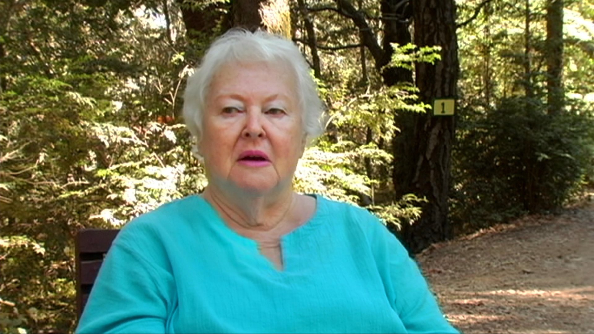 An older woman shares personal memories during a documentary-style interview, punctuated by archival footage of a man on a motorcycle. The setting transitions between a serene outdoor environment and historical personal imagery.