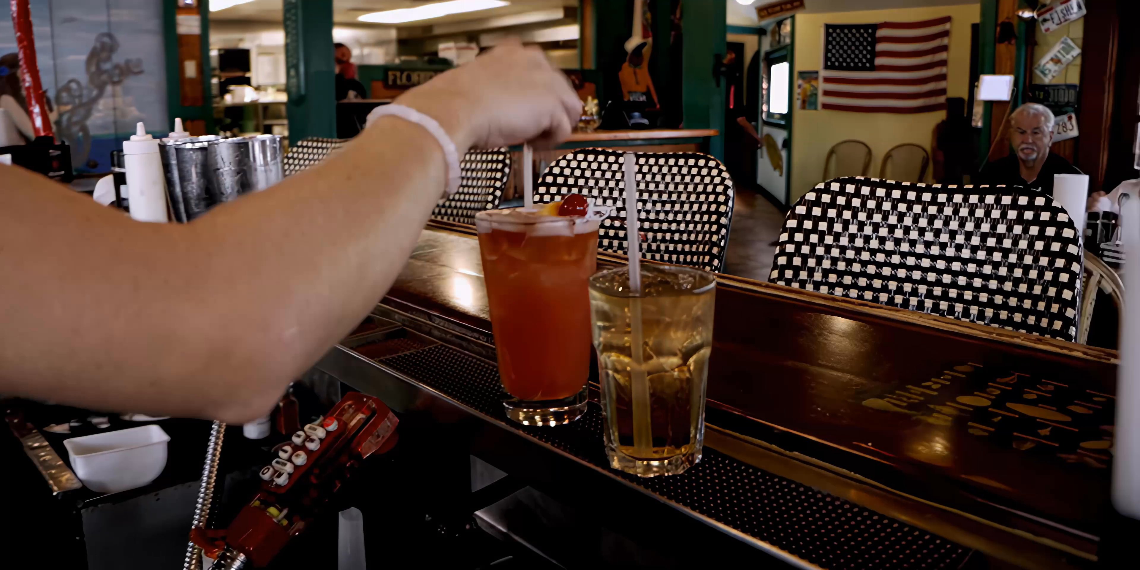A bartender expertly prepares and serves cocktails to patrons seated at a restaurant booth, emphasizing efficient professional service. The scene captures the transition from drink preparation behind the bar to delivering refreshments to customers in a relaxed dining atmosphere.