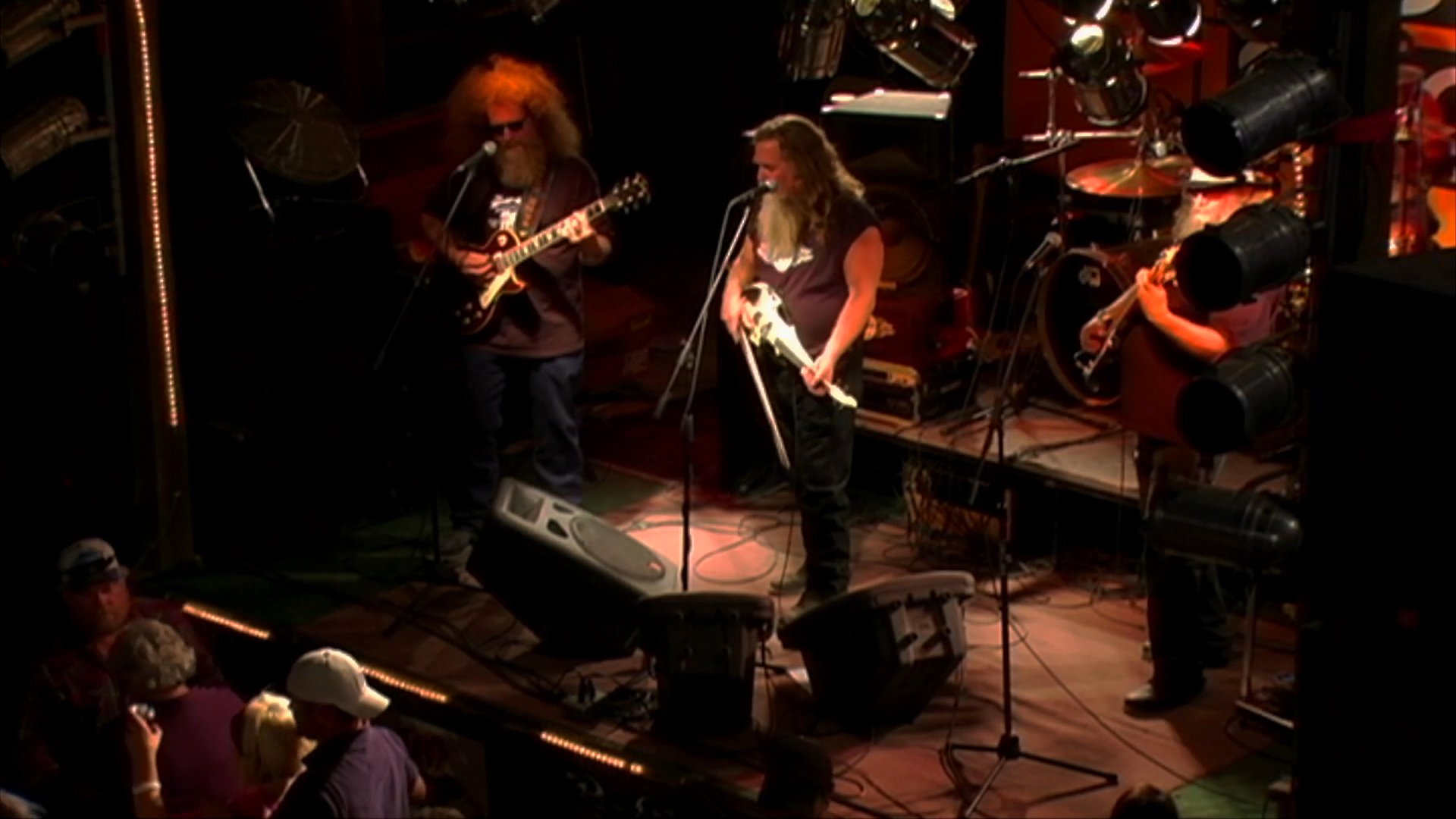 A rock band performs live on stage at a crowded music venue, featuring a long-haired lead singer and guitarists engaged in a musical set.