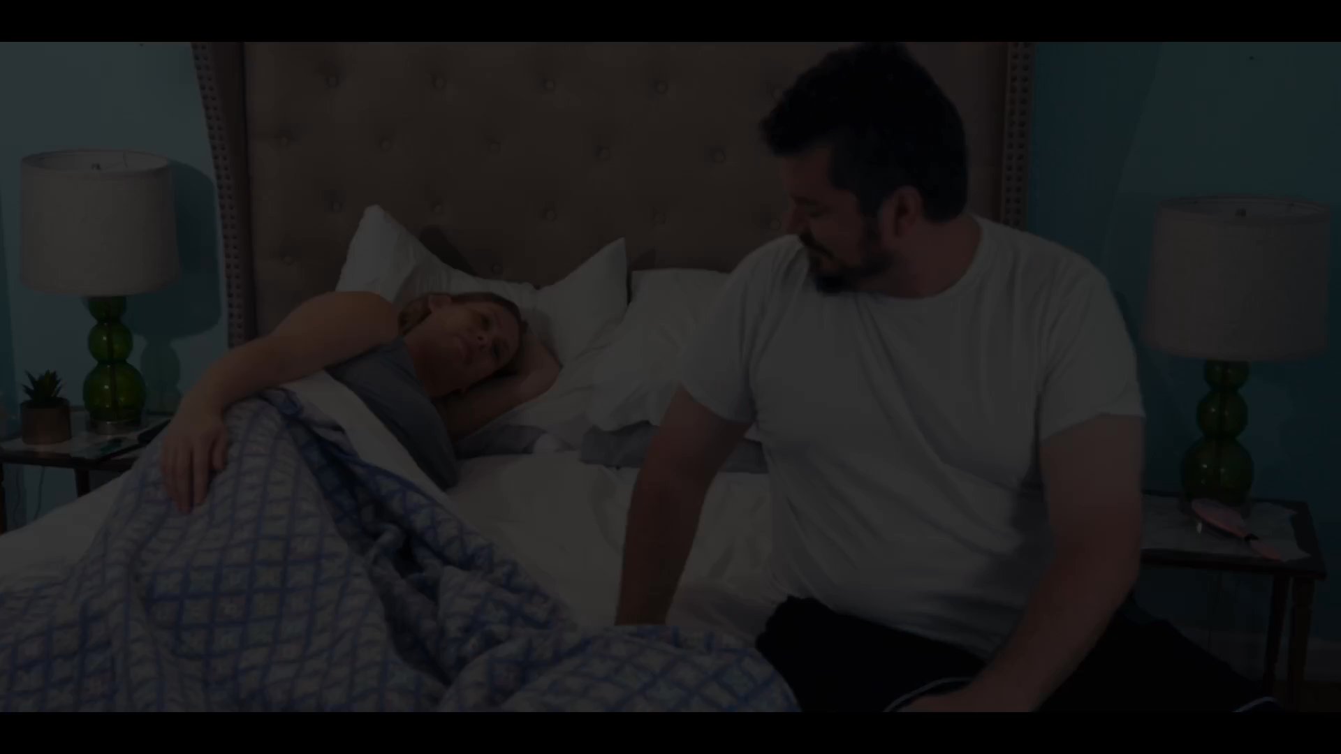 A man wakes up beside a woman in a bedroom before the camera transitions to an aerial view of a suburban neighborhood and follows a person walking toward a doorway. The sequence portrays a calm, domestic start to the day.