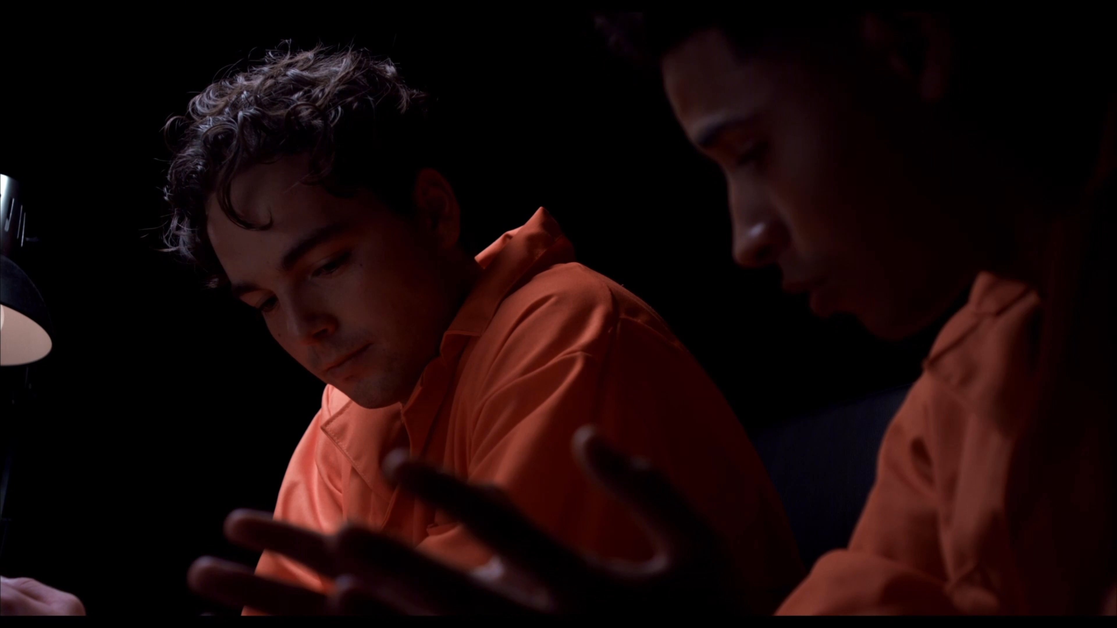 Two individuals dressed in orange prison jumpsuits engage in a serious, low-light conversation within a confined environment. The dialogue and body language suggest a high-stakes or tense interaction between the two participants.