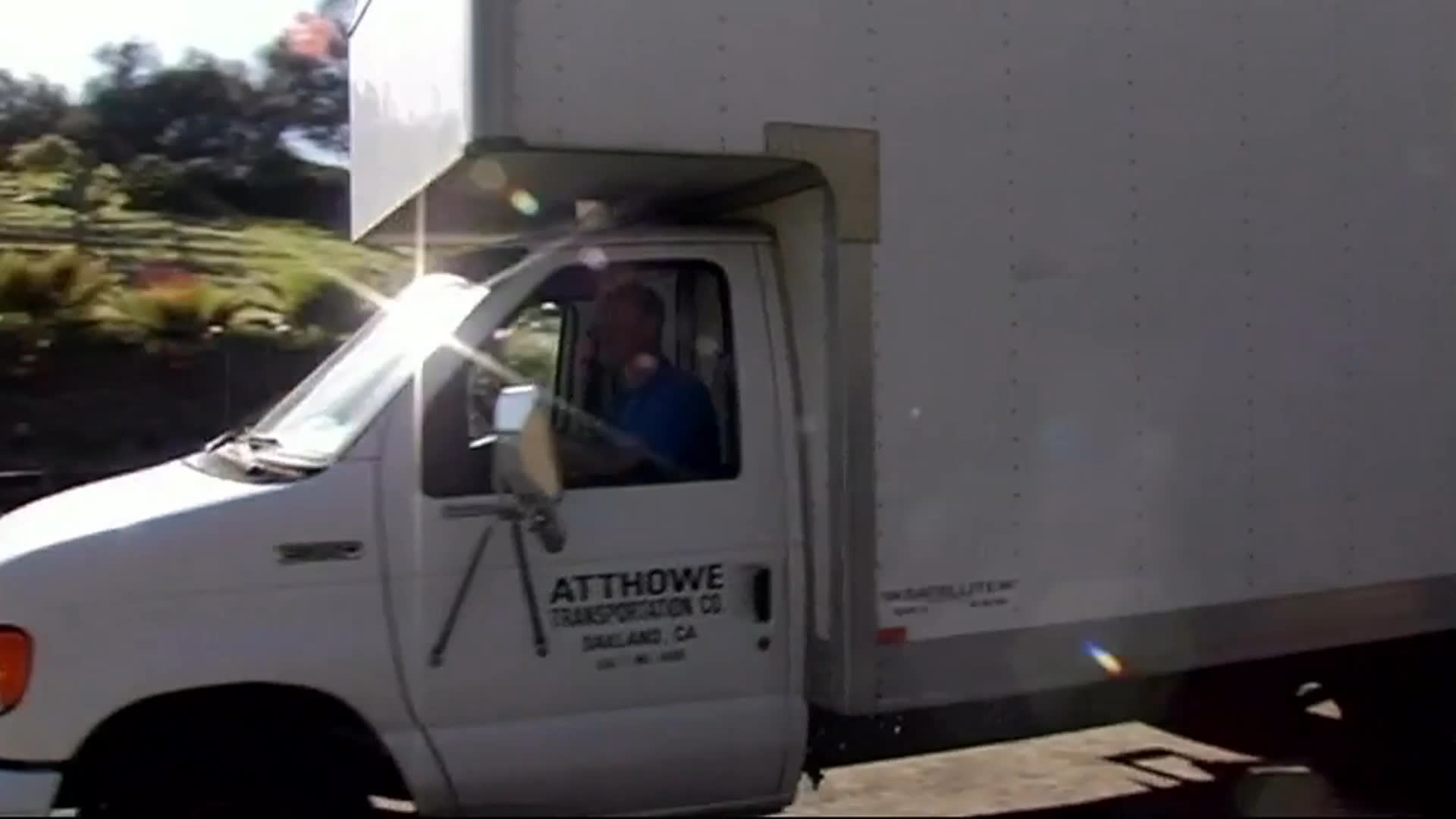 A white box truck pulls away from a driveway as a man in a green cap watches. After it leaves, he walks toward the camera across the sunlit lot.