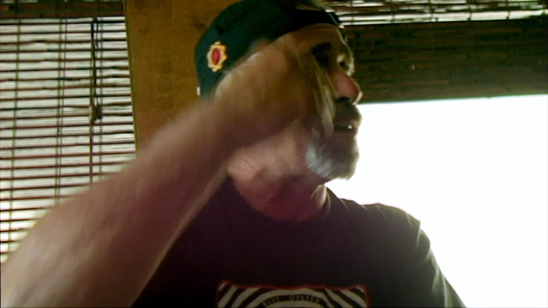 A man wearing a baseball cap speaks expressively with hand gestures while sitting indoors. He maintains a lighthearted, conversational tone throughout the interaction.