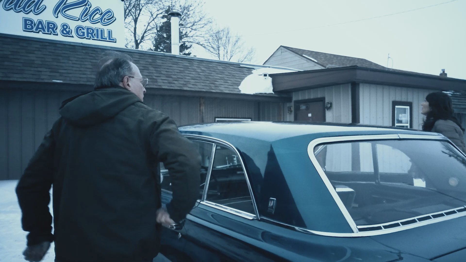 Individuals arrive in a vintage blue vehicle at a wintery restaurant location and proceed inside to socialize at the bar.