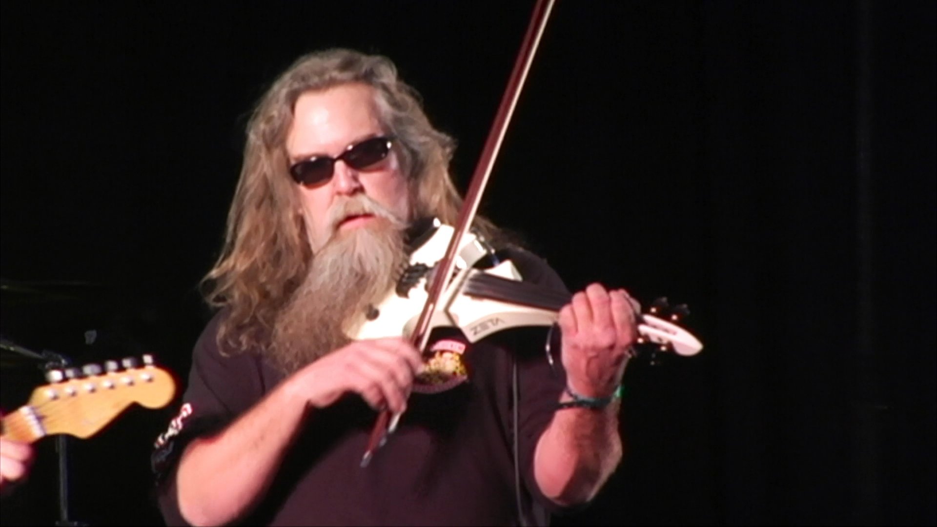 A long-haired, bearded violinist with sunglasses performs a lively solo on stage during a concert event. The musician plays an electric-style violin while accompanied by a band under stage lighting.