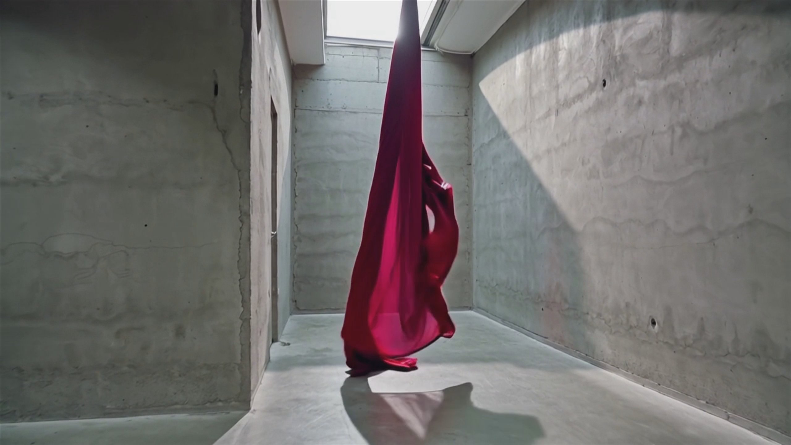 A crimson silk scarf drifts through a minimalist concrete gallery under a skylight.