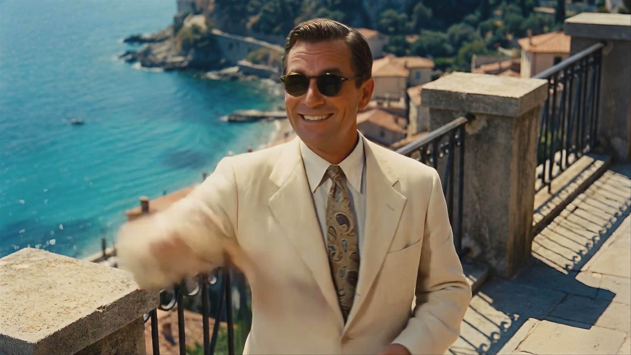 A man in a cream linen suit, patterned tie, and dark sunglasses stands on a stone balcony overlooking a Mediterranean coastal town with turquoise water. The shot is a medium close-up, generated using Kling AI Avatar v2 Standard at 1280x720 resolution. He is gesturing with his right hand.