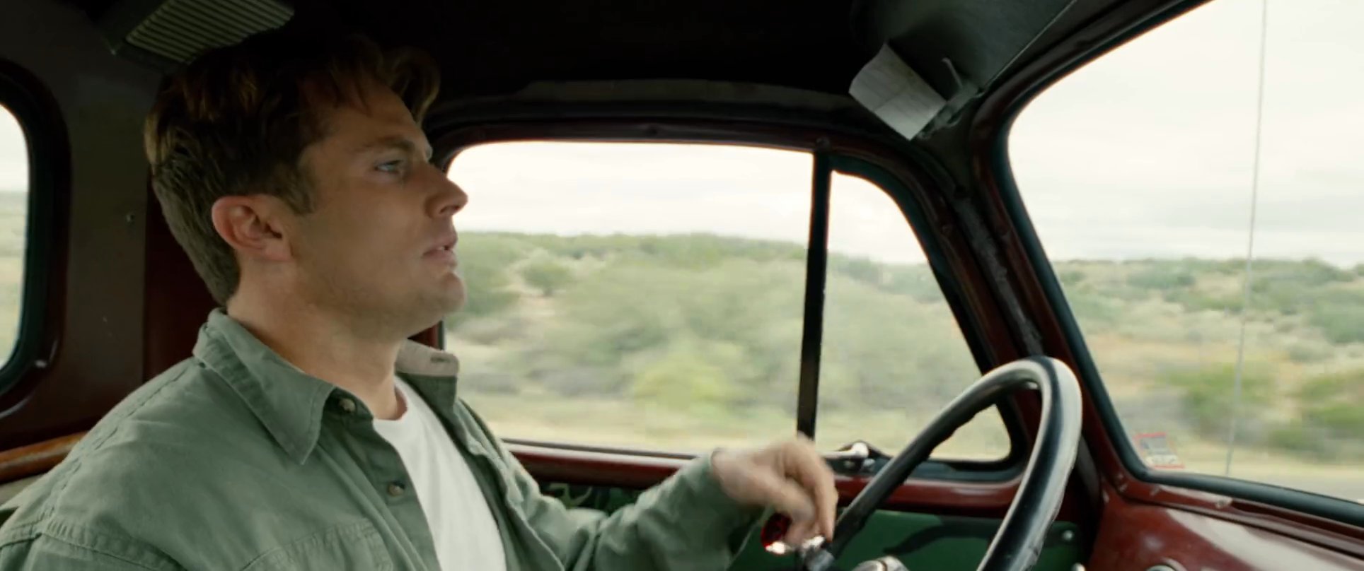 A man drives a vintage pickup truck through a desert landscape before arriving at a suburban residence to greet another man.