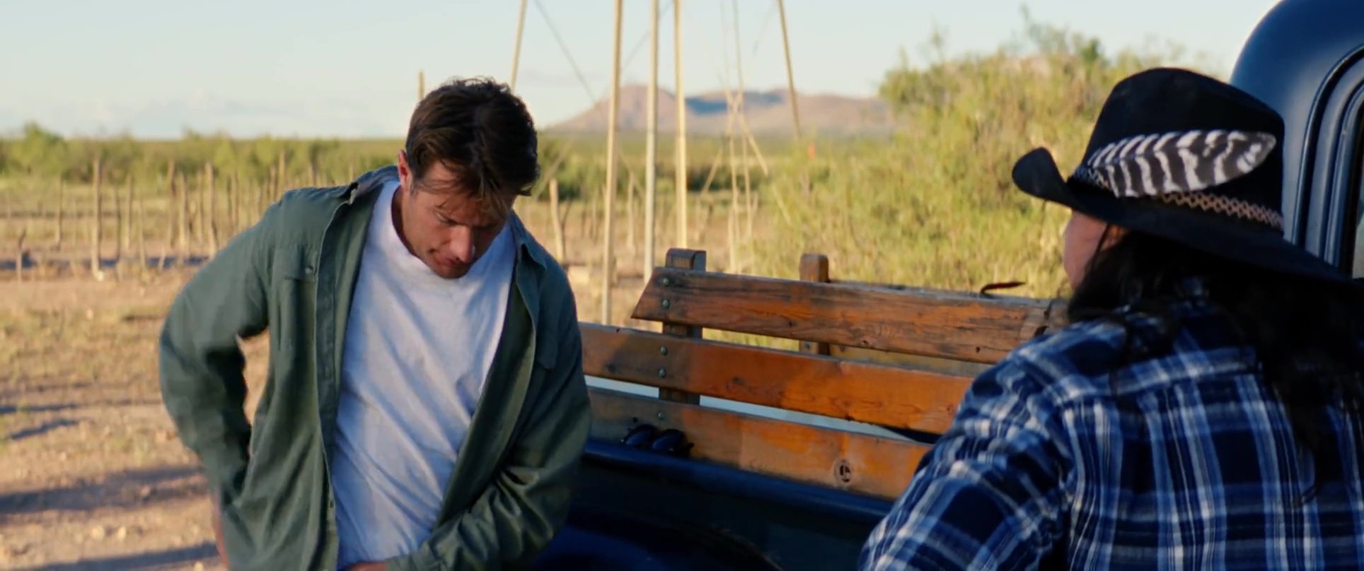 Two individuals conduct a financial transaction near a pickup truck in a rural desert setting. One person retrieves cash from a wallet to hand over to the other during an outdoor engagement.