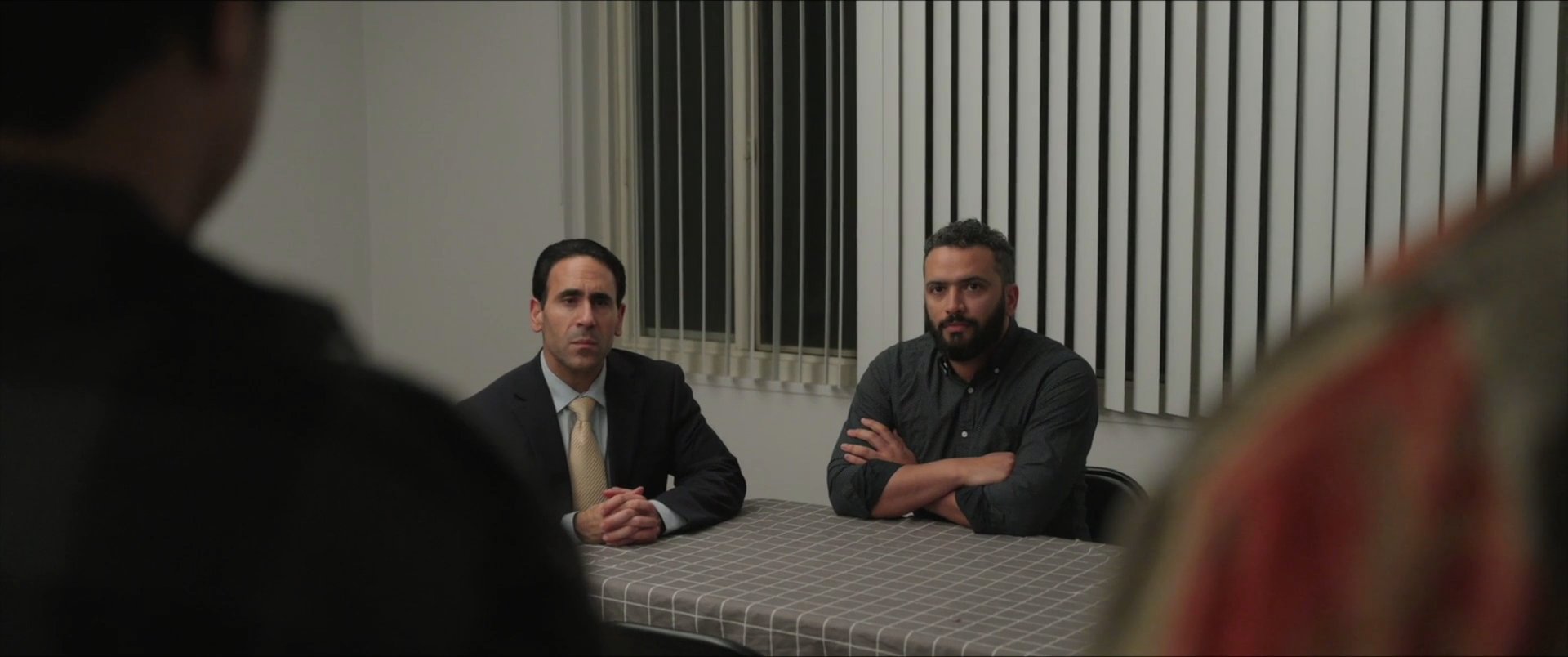 Two young men enter a professional office to engage in a formal meeting with two individuals seated at a table. The participants exchange serious dialogue across the table in a sterile, lit environment.