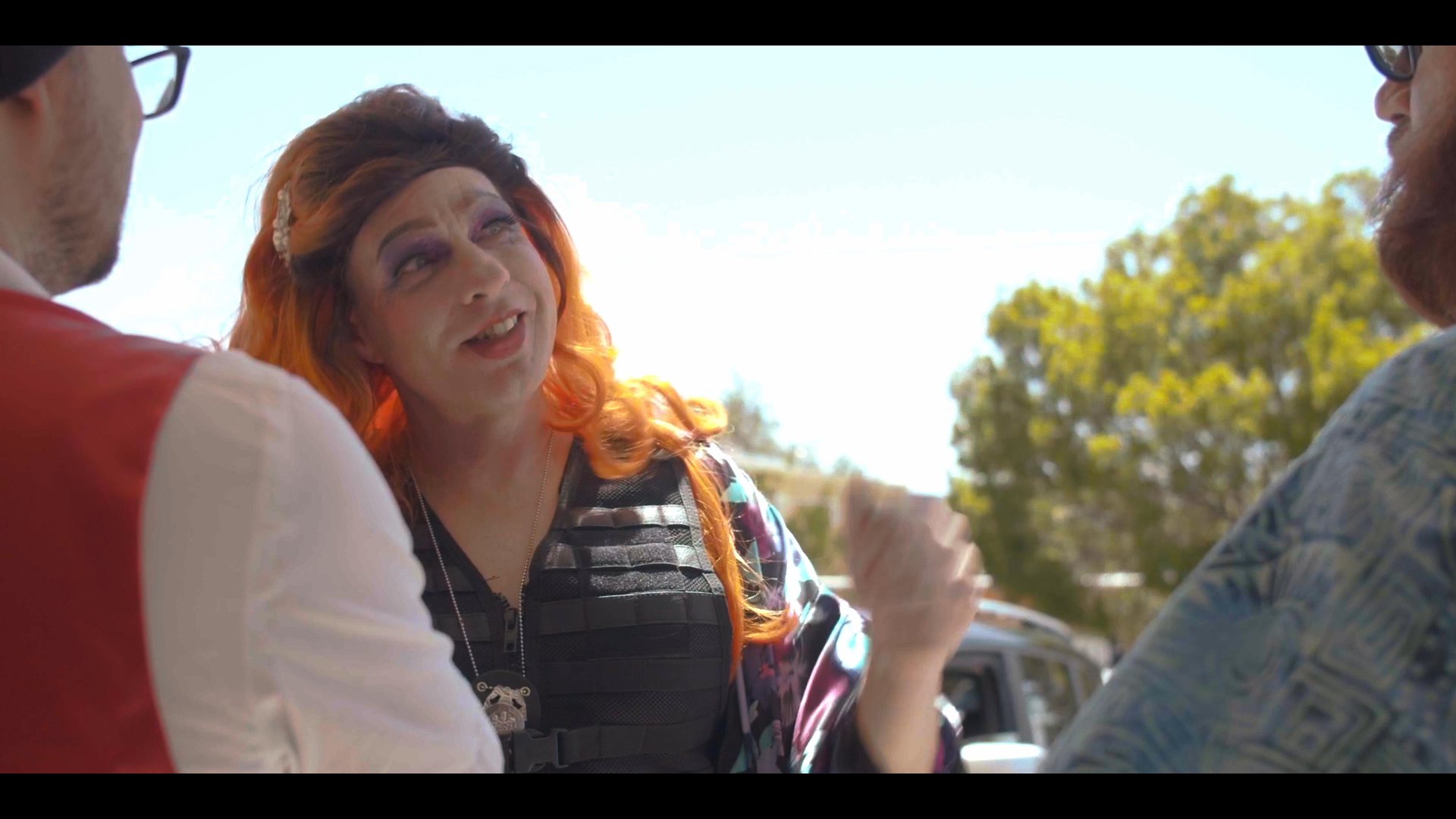 A drag performer with vibrant orange hair engages in a dramatic conversation with two men standing outside. The interaction features expressive gestures and theatrical styling set against an outdoor background.