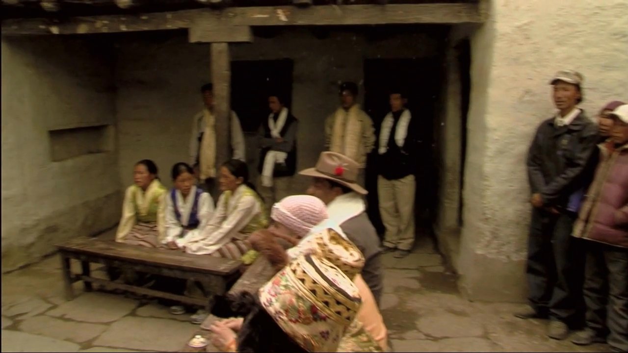 A teacher guides students through a lesson at a chalkboard before the community gathers for a formal, traditional ceremony. Attendees participate in cultural rituals involving white scarves in a rural mountain setting.