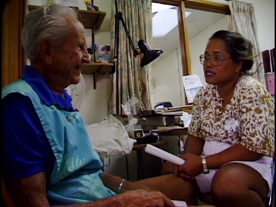 A healthcare provider engages in a supportive, personal dialogue with an elderly patient inside a clinical setting. The interaction maintains a warm tone, focusing on effective communication and patient comfort.