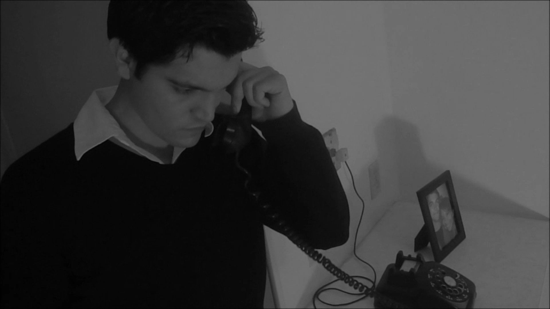 A man in a dimly lit room picks up a rotary telephone and engages in a somber, focused phone conversation.
