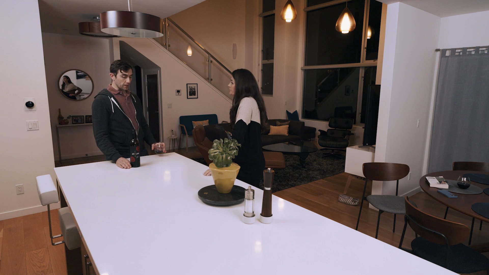 A man and woman engage in a serious, tense conversation while standing on opposite sides of a modern kitchen island. The man sips from a glass of wine as the two exchange words in a quiet living space.
