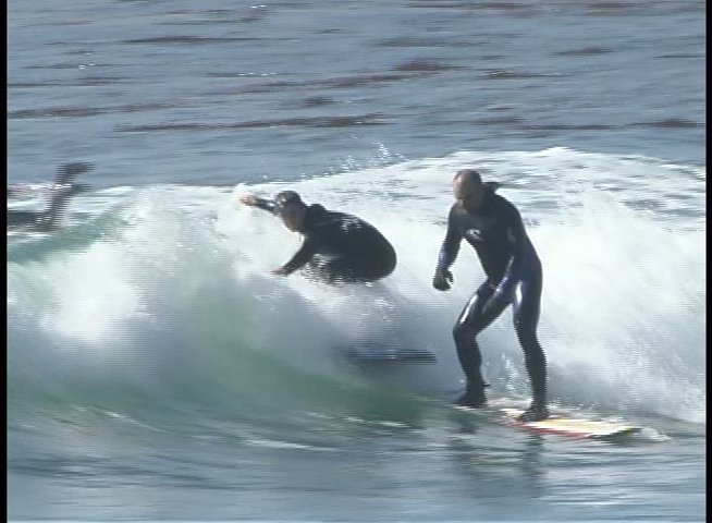 Multiple surfers in wetsuits navigate rolling waves on their surfboards in the ocean. The participants demonstrate balance and technique as they glide along the breaking waves.