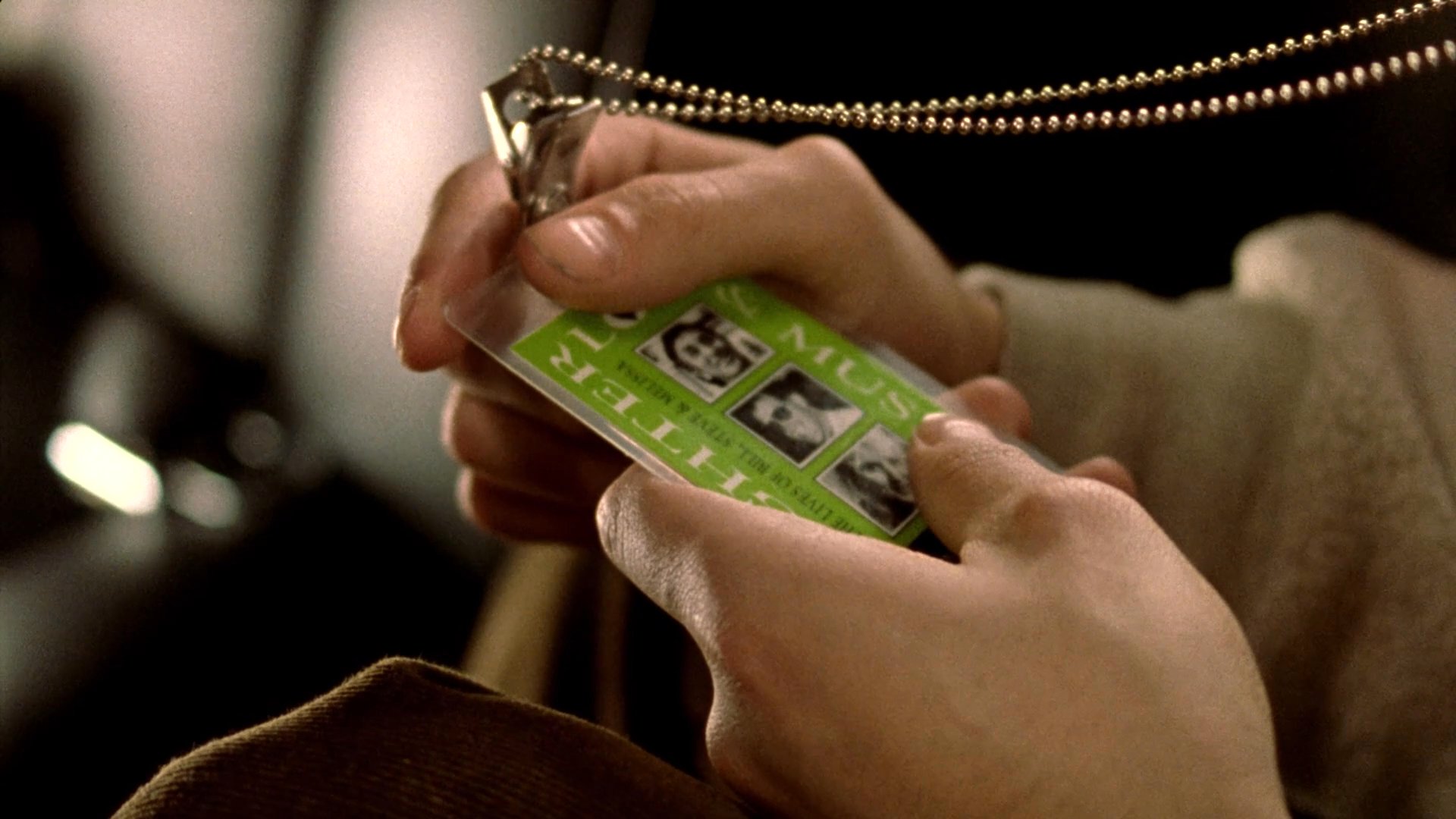 A passenger sits in the back of a limousine, closely examining an identification card hanging from a necklace while observing a miniature diorama of a traffic scene.