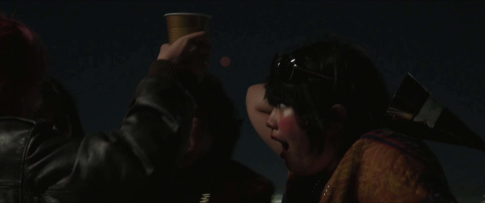 A group of friends celebrates New Year's Eve on a rooftop at night, sharing drinks, hugs, and kisses while watching fireworks. The atmosphere is festive and intimate as the group rings in the new year together.