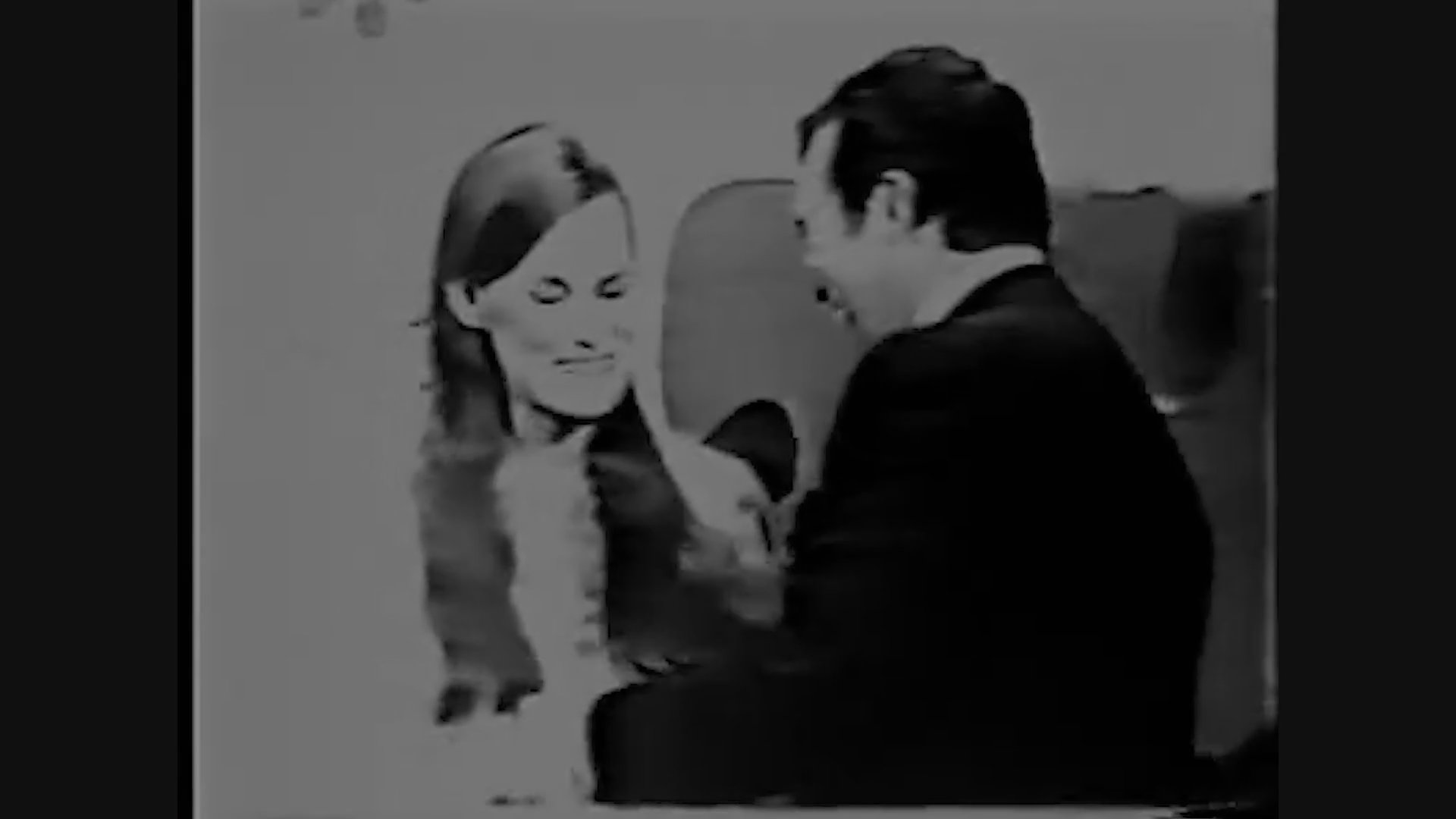 A television game show host conducts an interview with a female guest on a stylized, patterned studio set.