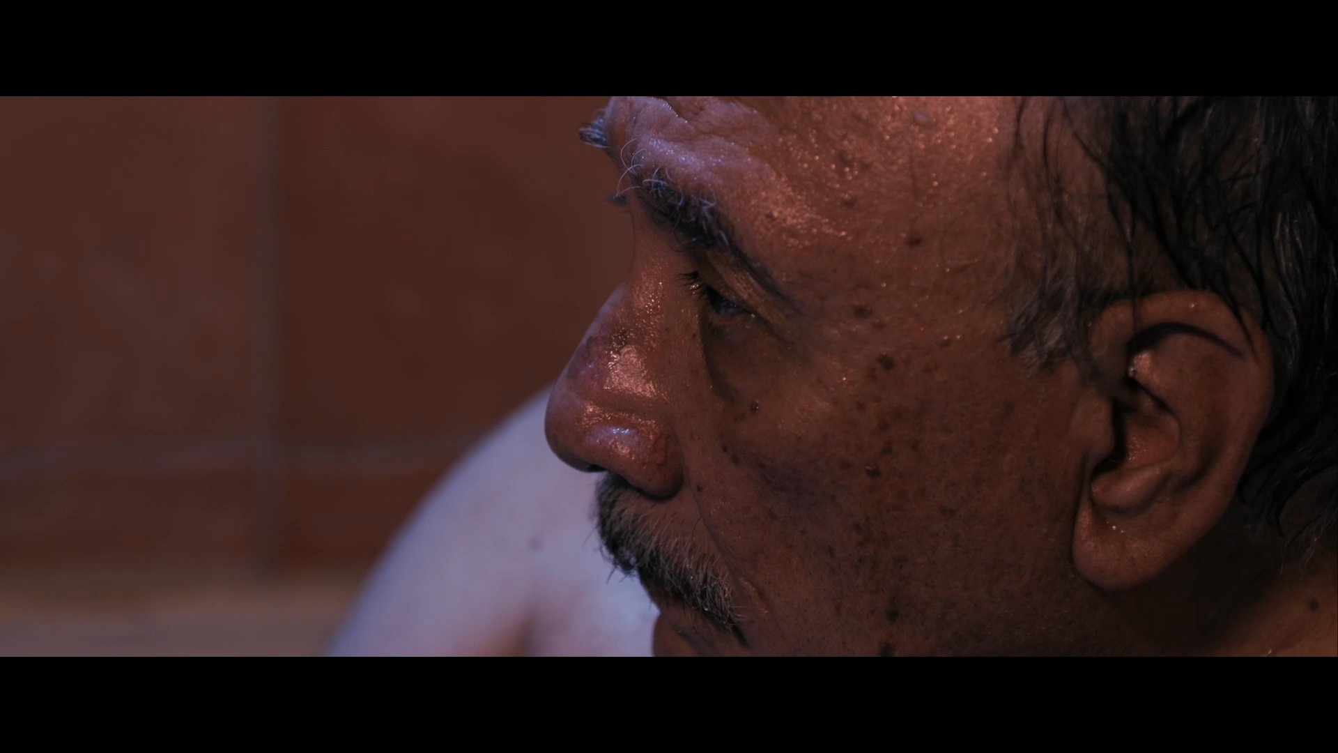An elderly man calls out for Carmen while sitting in a bathtub, visibly struggling to maintain composure or needing help.