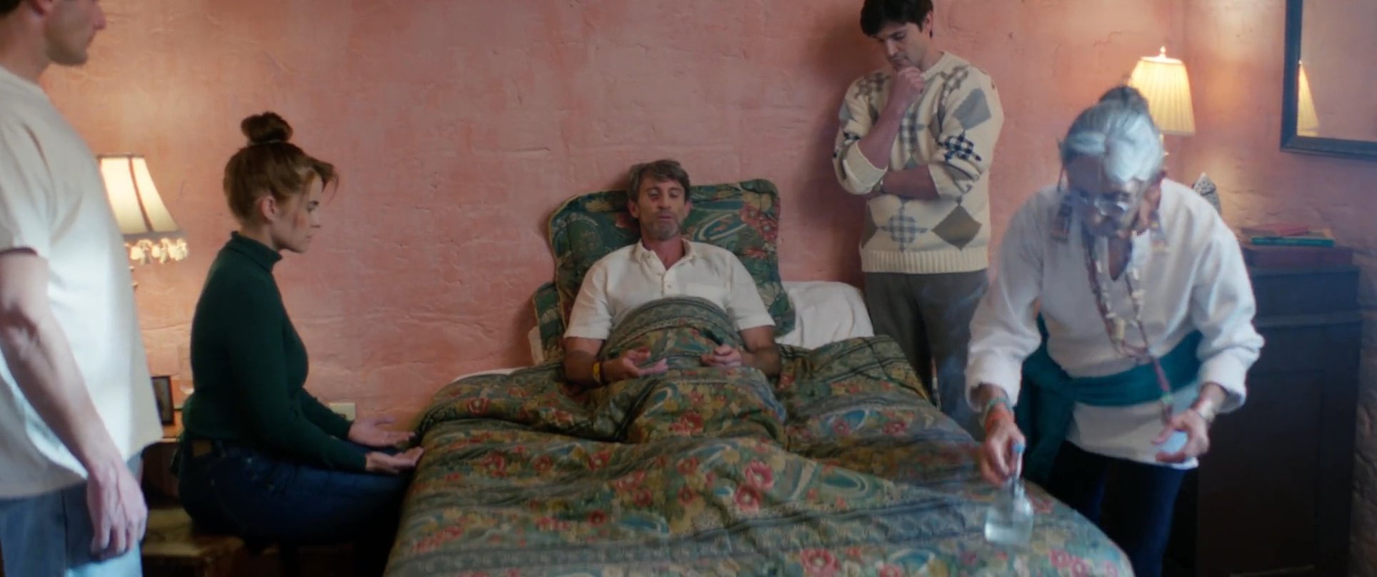 An elderly practitioner performs a spiritual healing ritual for a reclining man, while family members observe in a quiet room.
