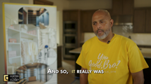 Sam Cotton wearing a yellow t-shirt, standing beside a display board of custom closet photos in a kitchen setting, from his Coffee in the Closet testimonial video