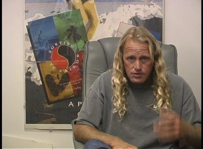 A man with long blonde hair discusses his experiences while sitting in a professional office setting. He gestures expressively as he recounts insights and stories related to surfing culture.