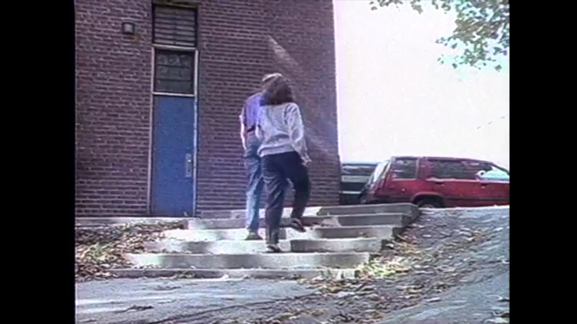 Two individuals walk together toward a brick building, ascending a set of concrete steps in an outdoor setting.