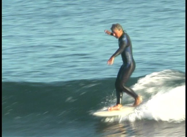 A man in a floral shirt provides commentary while footage showcases a surfer performing maneuvers on ocean waves. The segment emphasizes the skill and fluidity of surfing in a coastal environment.