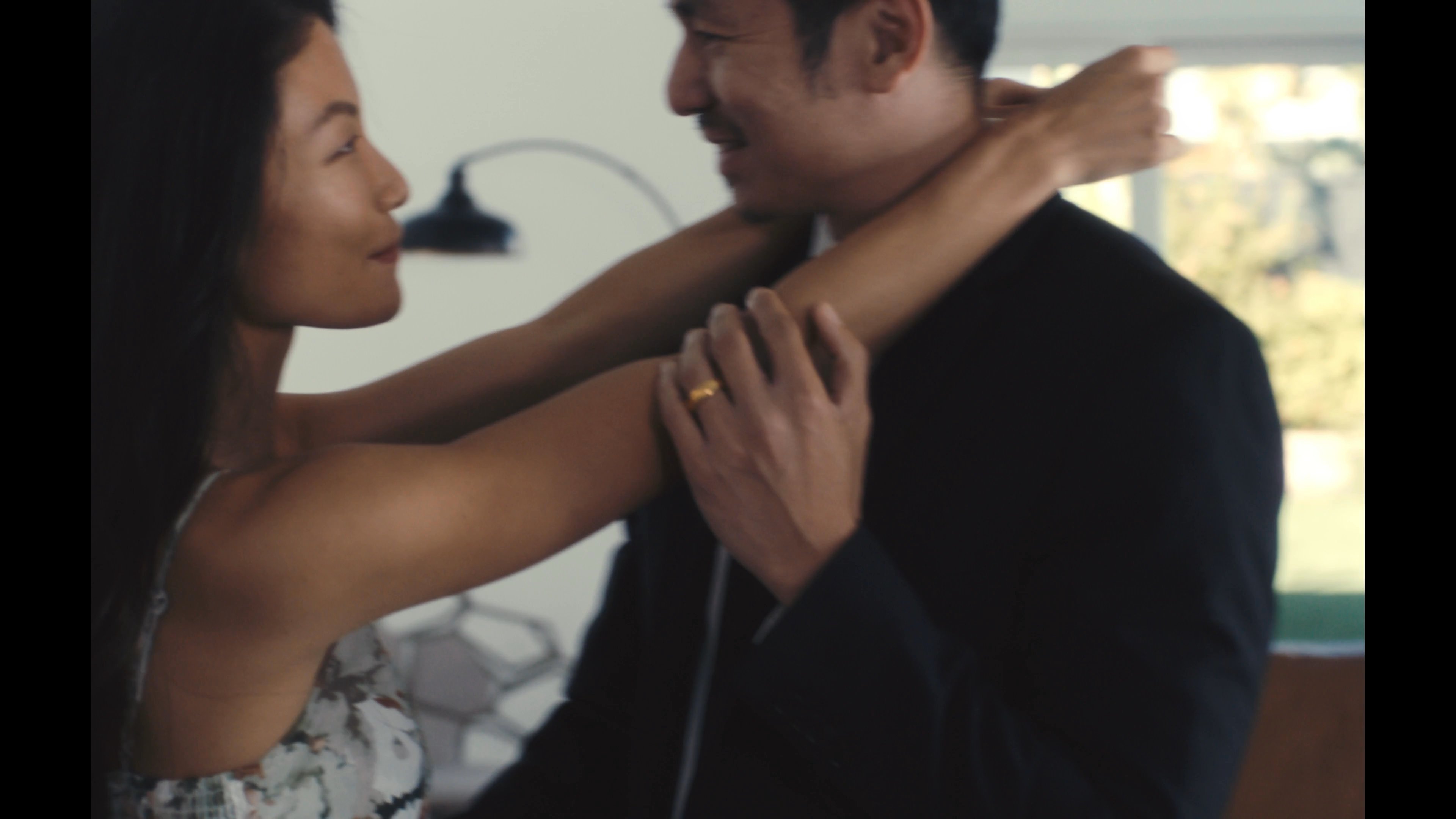 A well-dressed couple shares a romantic dance in a sunlit living room. The woman later looks toward the camera with a reflective expression.
