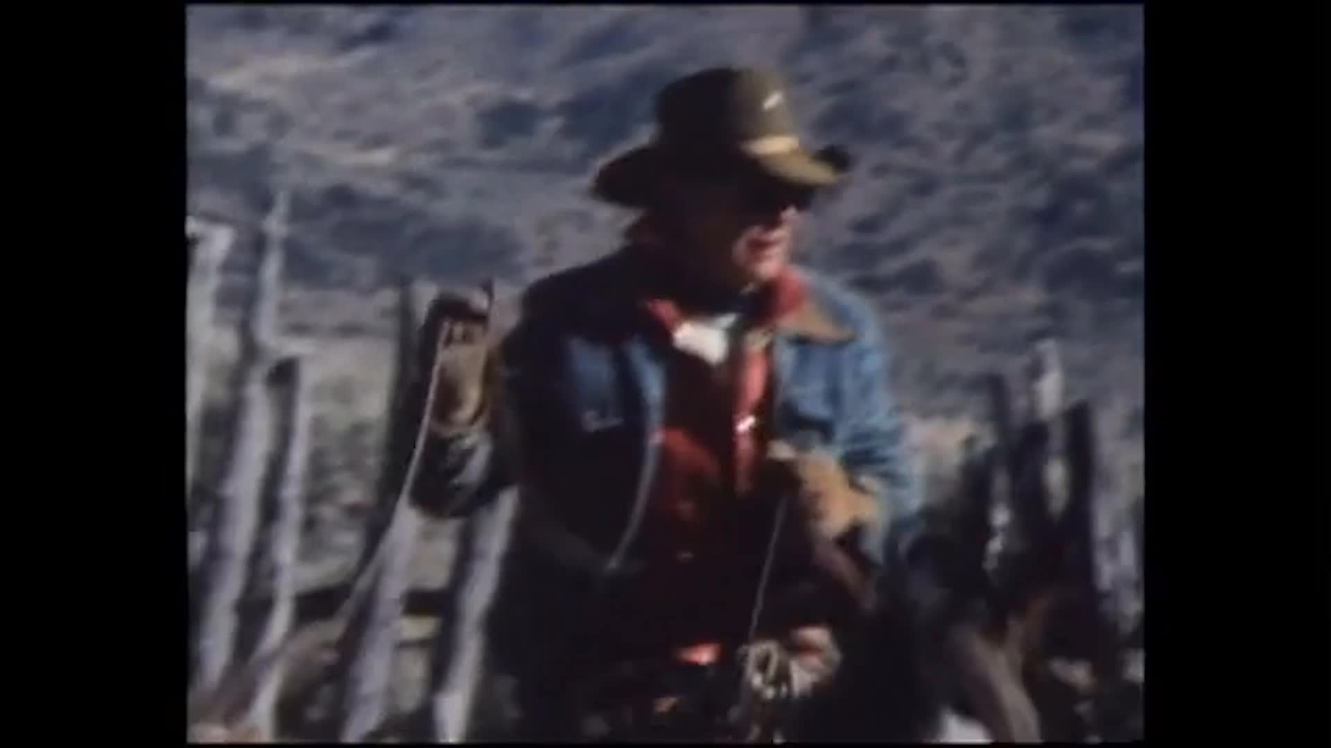 A rider on horseback guides cattle through a dusty corral. A cowboy-hatted rancher handles gear beside a weathered wooden structure, with mountain terrain in the background.